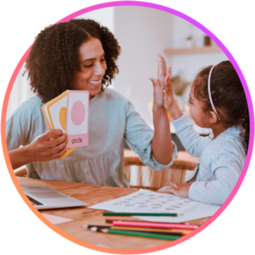 Teacher holding up keyword image cards and high-fiving a young student as they sit together at a work table